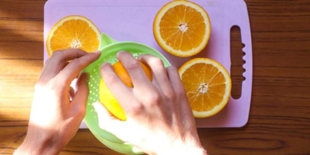 Mão espremendo laranja em um espremedor, apoiado em uma tábua roxa